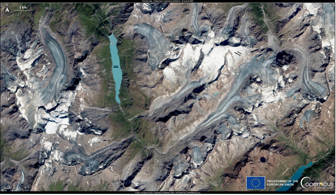 The Sentinel-2 satellite image, acquired on 22 August 2023, shows the Swiss Alps during a severe heatwave in Europe. The image vividly illustrates the impact of record-breaking temperatures, even at high altitudes, resulting in the dramatic melting of glaciers. The glaciers appear starkly exposed, having completely lost their winter ice cover, and many of the usually snow-covered peaks are barren and rocky. The image includes labelled glaciers such as "Glacier de Corbassière" and "Glacier du Mont Durand," with a scale bar indicating 1 km in the top left corner. The bottom right corner features logos for the European Union and Copernicus.