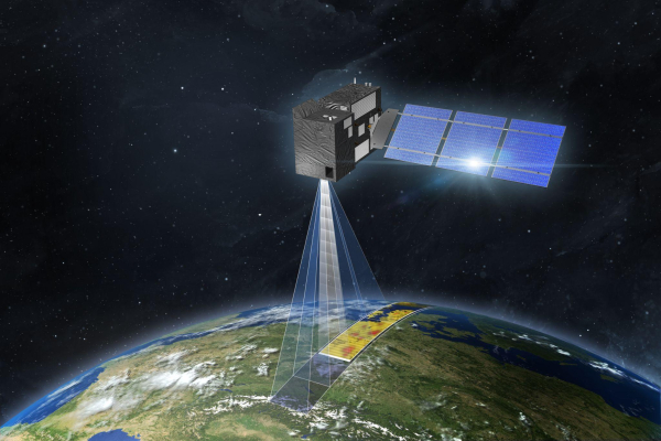 The image is an artist’s impression of a CO2M satellite in space. The satellite has a rectangular body with solar panels extending from one side. It is positioned above Earth, with a visible beam projecting downward, indicating the satellite's monitoring capabilities. The Earth's surface below shows a detailed section of land with varying colours, likely representing data collected by the satellite. The background features the dark expanse of space dotted with stars. This satellite image is credited to OHB 