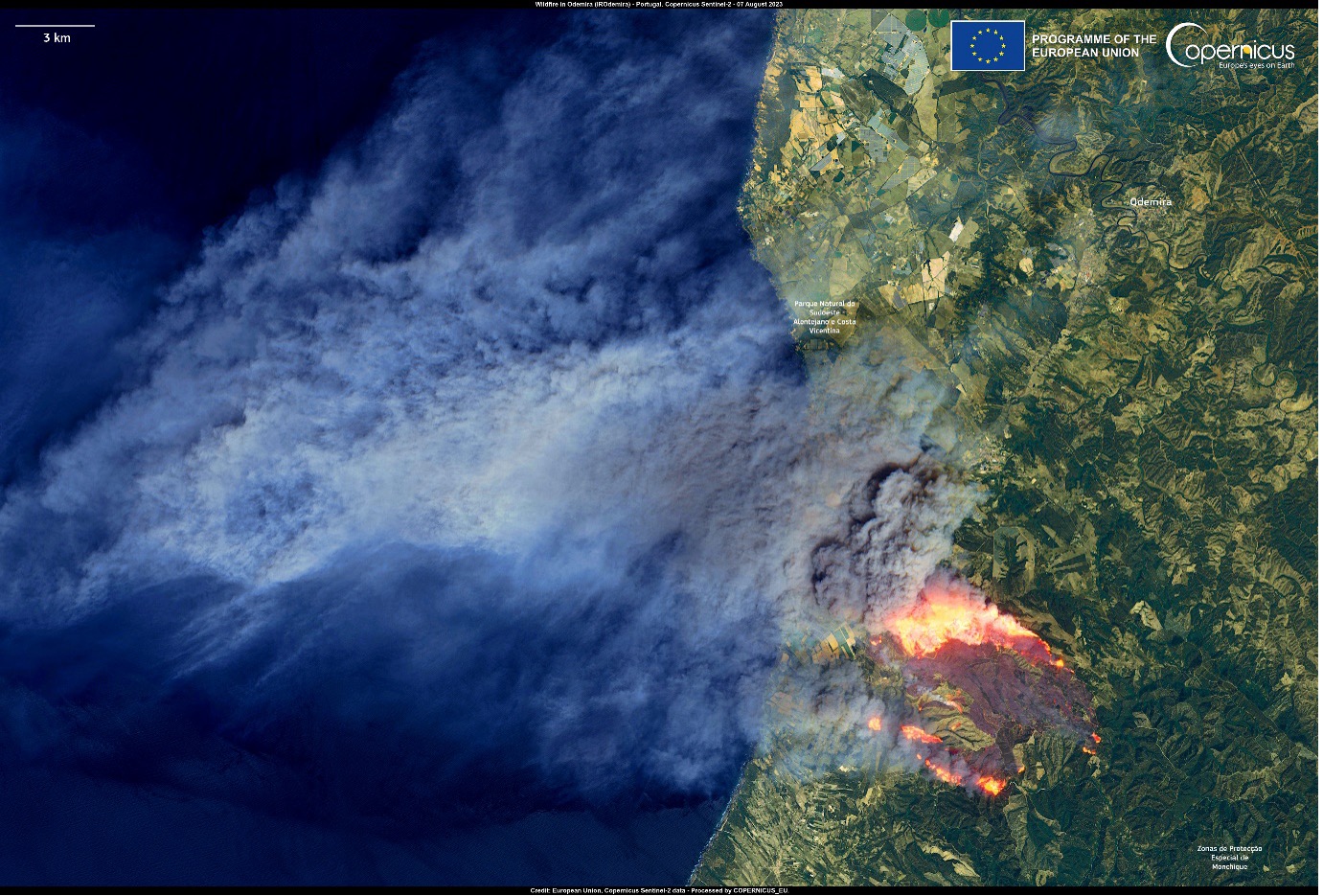  The image captures a severe wildfire in Portugal, primarily affecting the Odemira municipality in the Alentejo region. The satellite image shows bright orange and red fire fronts with a large plume of smoke extending northwest over the Atlantic Ocean. The land surrounding the fire appears dry and parched, with the fire spreading rapidly through the area. The image is overlaid with labels for locations such as "Parque Natural do Sudoeste Alentejano e Costa Vicentina" and "Zonas de Perceção Incêndio Monchique," and includes a scale bar indicating 3 km. The top right corner displays logos for the European Union and Copernicus.